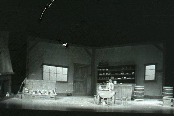 A dimly lit room with rustic furniture, including a table, chairs, and wooden barrels. Shelves with dishes are visible in the background. The setting resembles an old country inn or tavern.
