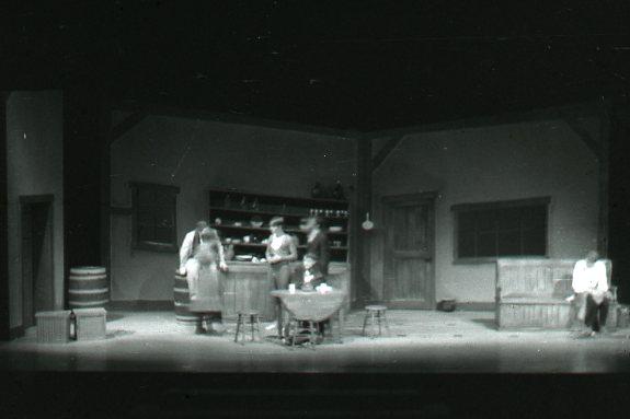 A black and white photo of a stage set depicting a rustic room. There are five people; four gathered around a table in the center, and one seated on a bench to the right. Shelves with bottles and barrels decorate the background.