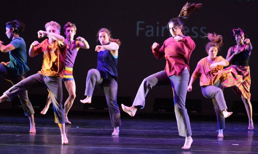 A group of seven dancers perform on stage, each in mid-motion with one leg raised. They wear colorful, casual attire and display various dance poses. The dark background has faint, partially visible text.