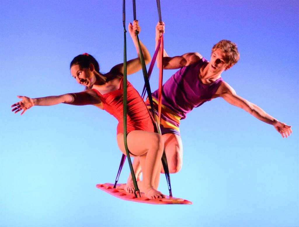 Two performers in colorful outfits balance on a trapeze against a gradient blue background. They are facing opposite directions, each with one arm extended, appearing joyful and energetic.