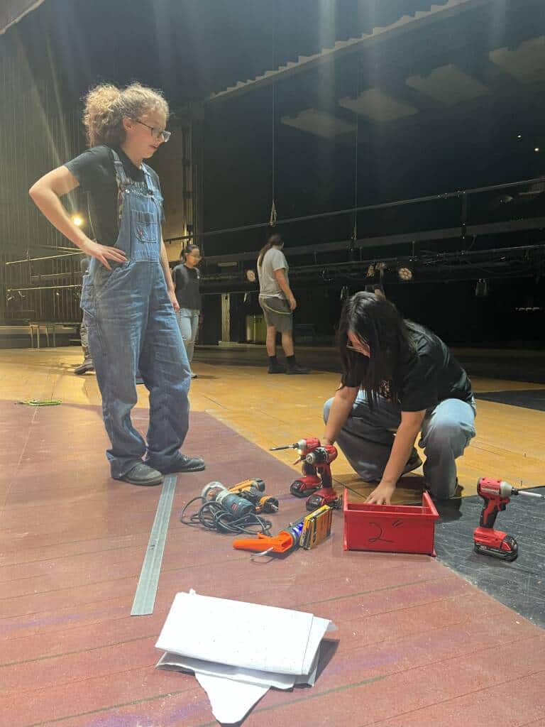 Two people work on a stage; one stands wearing overalls while the other kneels near tools and a red box. In the background, others are also working under stage lighting. A tape measure and paper sit on the floor.