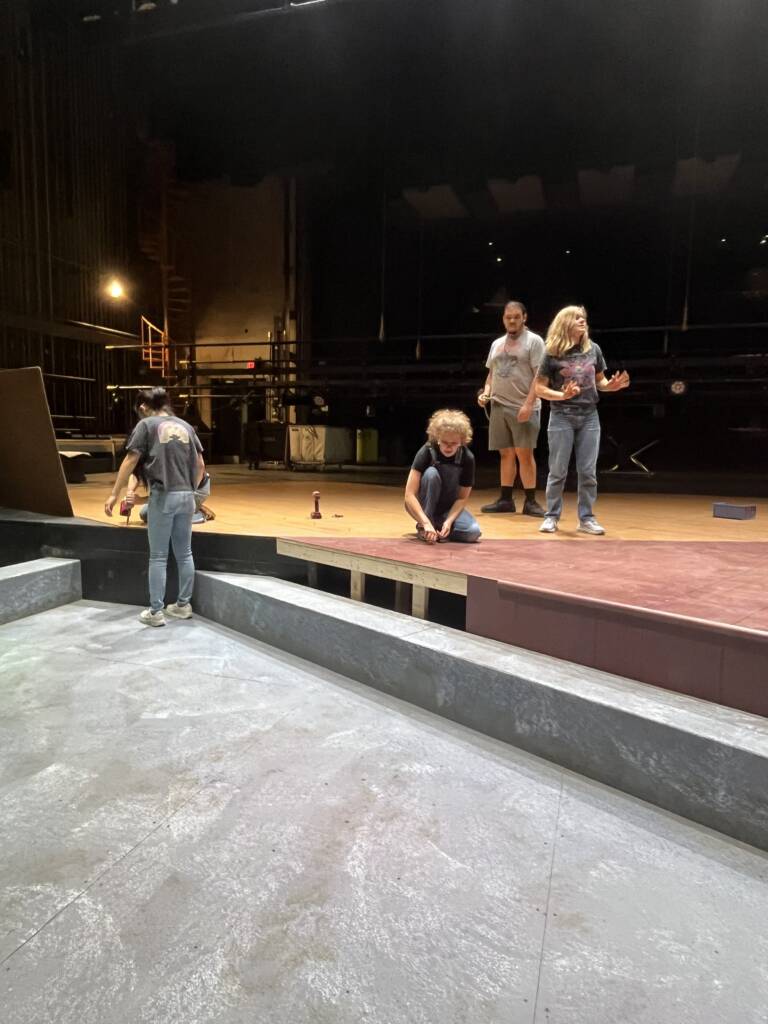 Four people work on a theater stage set. Two kneel on the floor using tools, while two others stand talking. The background is dark with visible stage equipment and lighting.