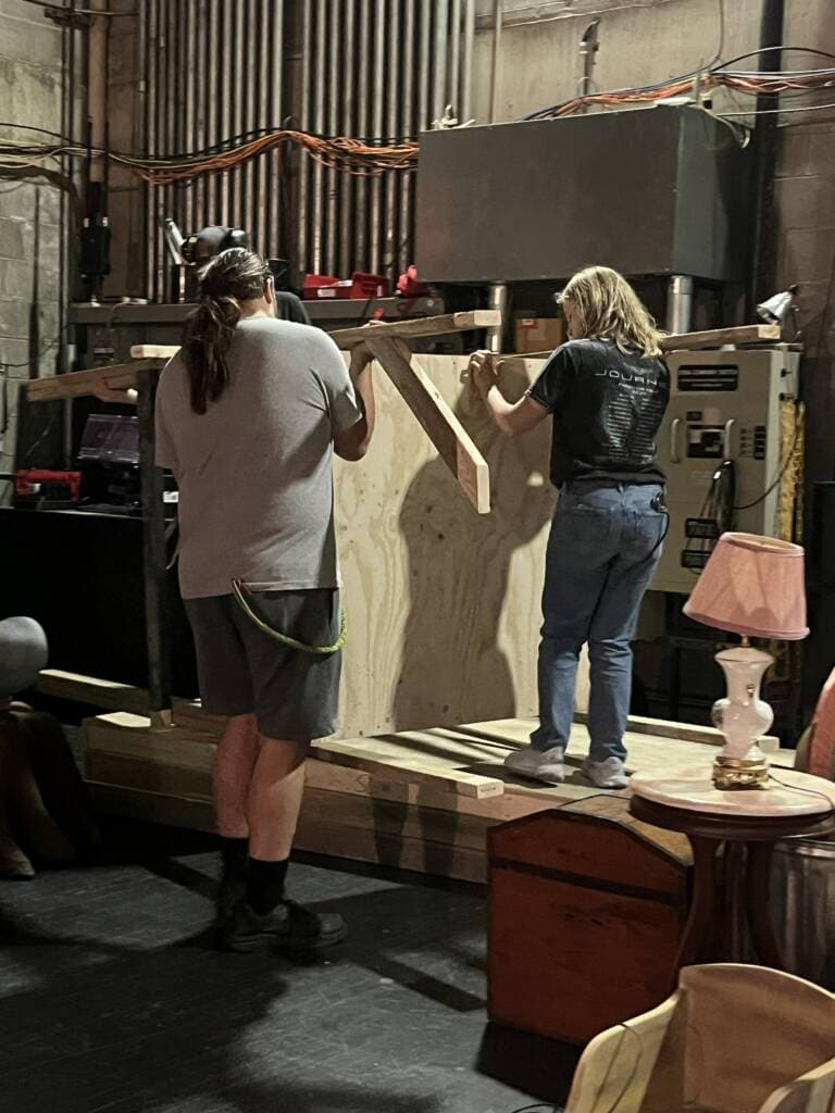 Two people, seen from behind, are assembling a large wooden structure indoors. They are surrounded by various stage props, including a lamp, a chair, and some cables on the wall in a backstage setting.