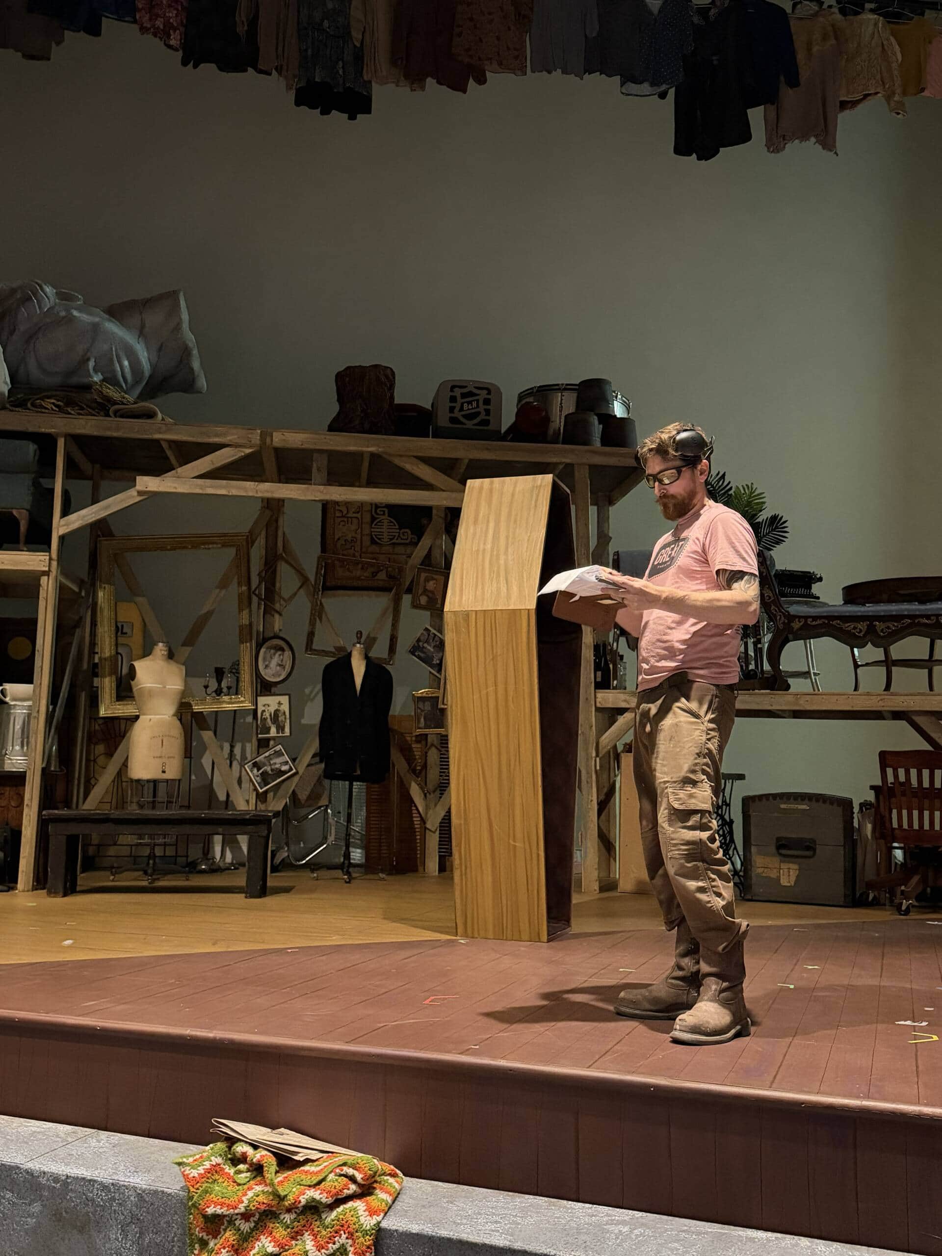 A man in a pink shirt and cargo pants stands on a stage reading from a script. Behind him is a wooden structure with vintage items, mannequins, and hanging clothes, creating a theatrical set.