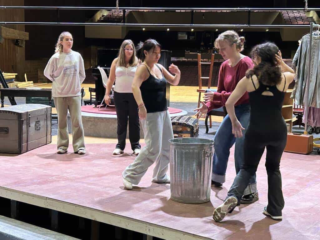 Five women rehearse on a theater stage. One woman stands near a metal trash can while others interact around her. Stage props and costumes are visible in the background under stage lights.