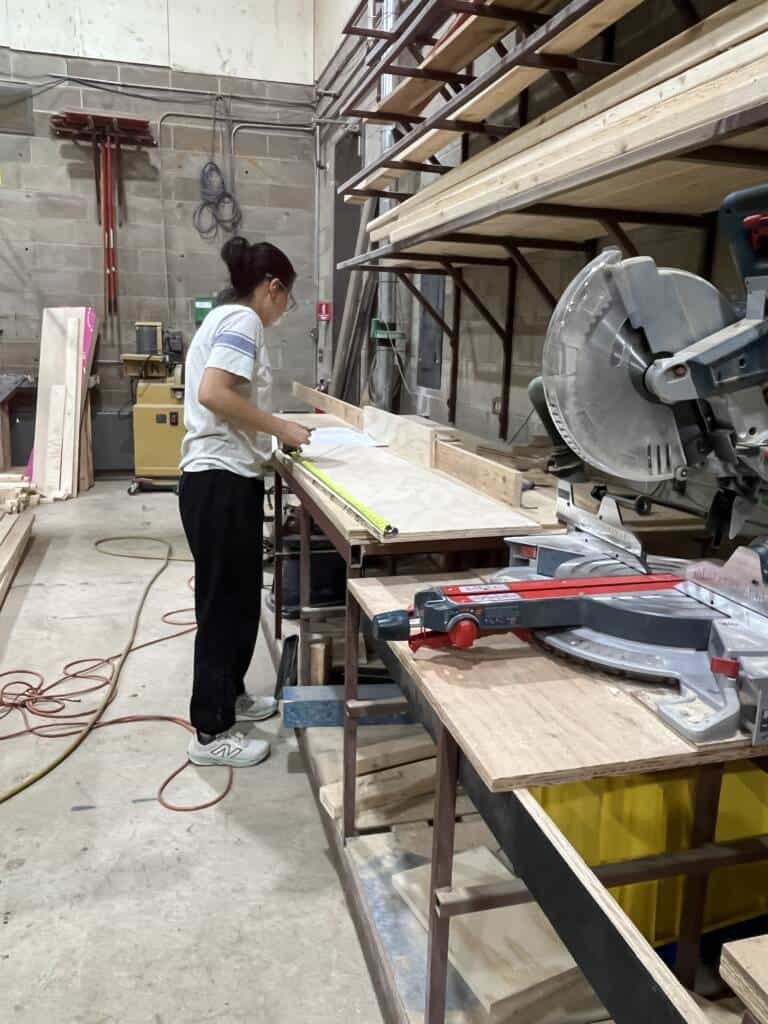 A person measures a piece of wood on a workbench in a workshop. Nearby are a circular saw, shelves with wood planks, and various tools. The scene is well-lit and organized.