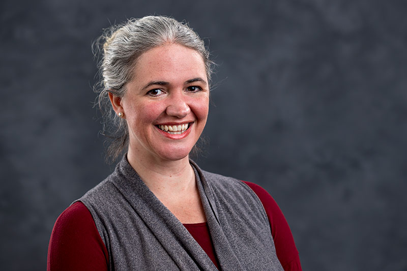 A woman with gray hair tied back, smiling at the camera. She is wearing a gray sleeveless vest over a maroon top. The background is a plain, dark, and textured backdrop.