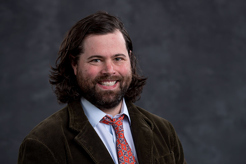 A smiling man with shoulder-length brown hair and a beard is wearing a striped shirt, red patterned tie, and a brown corduroy blazer. The background is a dark, textured gray.