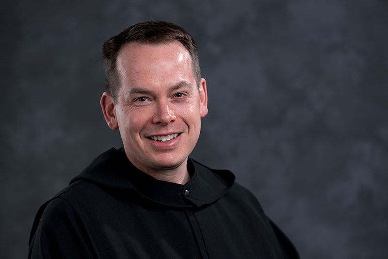 A man with short dark hair, wearing a black robe, smiles against a mottled gray background.