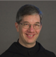 A man with short gray hair, wearing glasses, smiles. He is dressed in a black outfit against a dark gray background.
