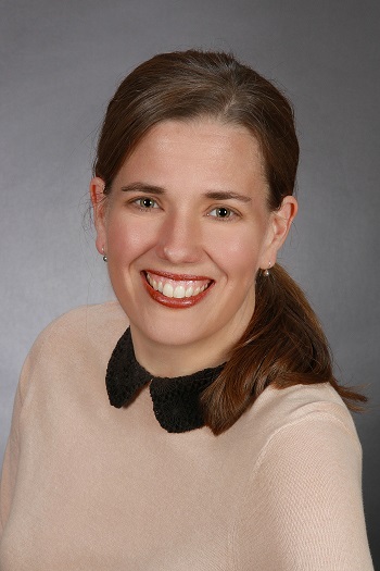 A smiling woman with light skin and brown hair tied back in a low ponytail. She is wearing a beige top with a black collar. The background is solid gray.