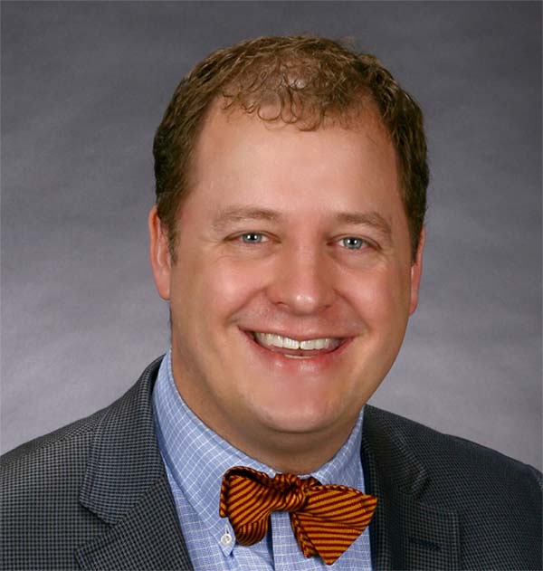 A man with short curly hair is smiling. He is wearing a blue checkered shirt, an orange and black striped bow tie, and a dark gray blazer. The background is a plain dark gray.