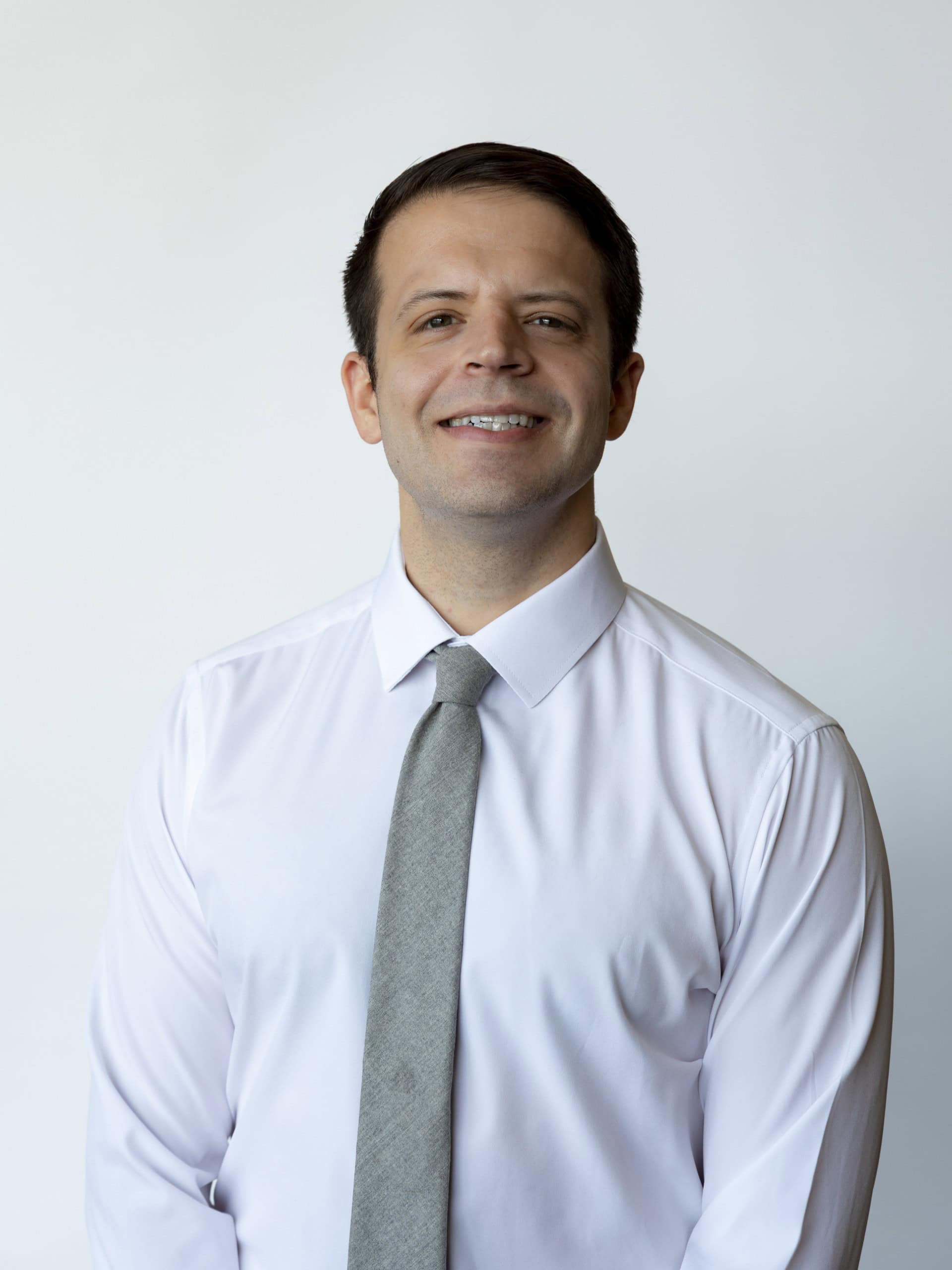 A man wearing a white dress shirt and a gray tie stands smiling against a plain white background.