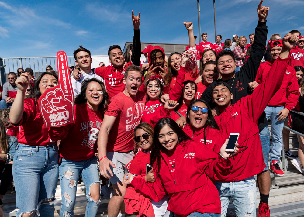 A large group of enthusiastic college students, wearing red St. John's University attire, gather on bleachers. Some hold foam fingers and signs, while others take selfies and cheer, showing school spirit at an outdoor event under a clear blue sky.