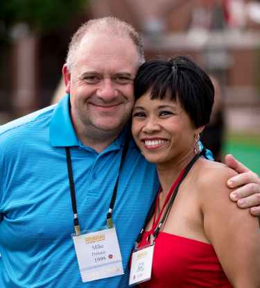 A smiling man in a blue polo shirt and a woman in a red strapless dress, both wearing name tags, pose together outdoors. The man has his arm around the woman, and they are standing against a blurred green and brown background.