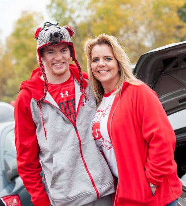 A young man and woman smiling at the camera. The man is wearing a red and gray hoodie with a playful animal-themed knit hat, and the woman is wearing a red jacket over a white shirt. They are standing in front of an open car trunk, with trees in the background.