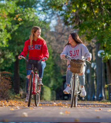Two young women ride bicycles down a tree-lined path. One wears a red "St. Ben's" hoodie, while the other has a gray "SJU" hoodie. They are smiling and appear to be engaged in conversation. The ground is scattered with fallen leaves, and the sun is shining.