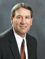 A man wearing a dark suit, white shirt, and maroon tie is posing for a portrait against a plain, dark background. He has short, dark hair with some gray and is smiling slightly at the camera.