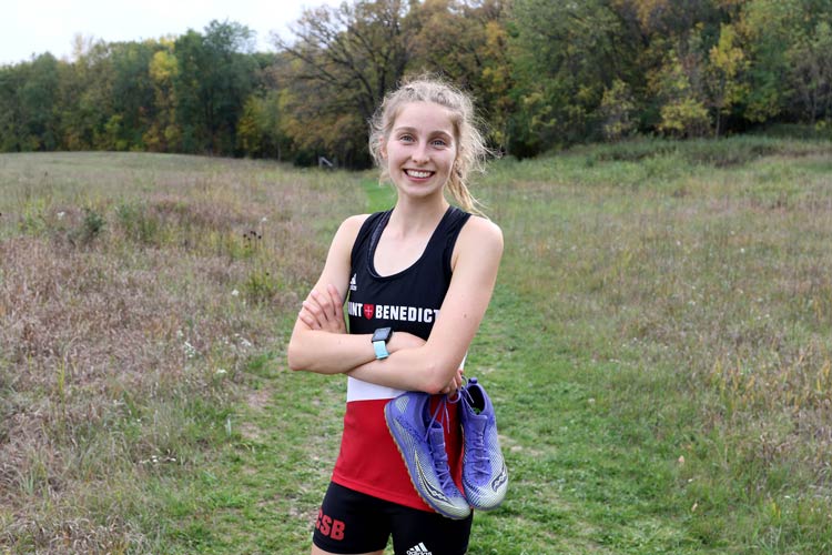 A smiling young woman stands in a grassy field with trees in the background. She is wearing a black, red, and white athletic uniform and has a pair of purple running shoes slung over her shoulder. Her arms are crossed, and she looks ready for a run.