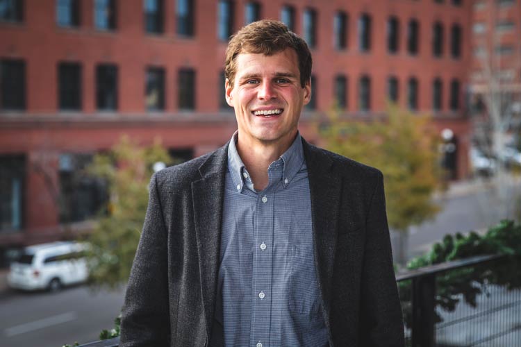 A man with short brown hair stands outside, smiling. He is wearing a dark blazer over a light blue button-up shirt. The background features a brick building and some trees, and the scene appears to be taken on a city street.