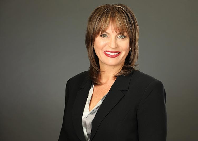 A woman with shoulder-length brown hair and bangs is smiling at the camera. She is wearing a black blazer over a silver blouse. The background is a plain, dark gray.