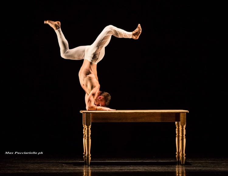 A male acrobat performs a handstand on a wooden table against a black background. He is balanced on his forearms, with his body extended upward and his legs gracefully bent. The acrobat is dressed in white attire.