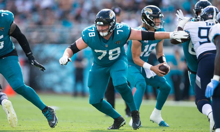 A football player in a teal uniform with the number 78 blocks an opponent, while the quarterback, also in a teal uniform, looks to pass the ball. Other players are visible in the background on a grassy field during a game.