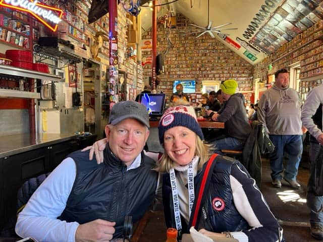 A smiling couple dressed warmly in black vests and hats sit together at a cozy, busy pub. The pub's walls are covered with various memorabilia, and other patrons can be seen in the background. The atmosphere appears lively and casual.