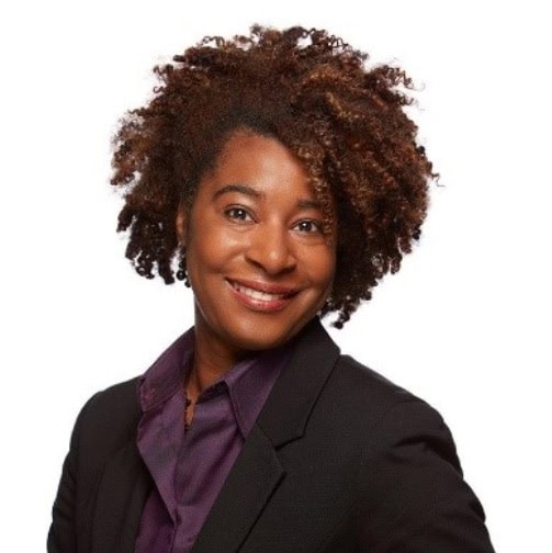 Portrait of a smiling individual with curly, dark brown hair. They are wearing a dark blazer over a purple shirt, standing against a plain white background.