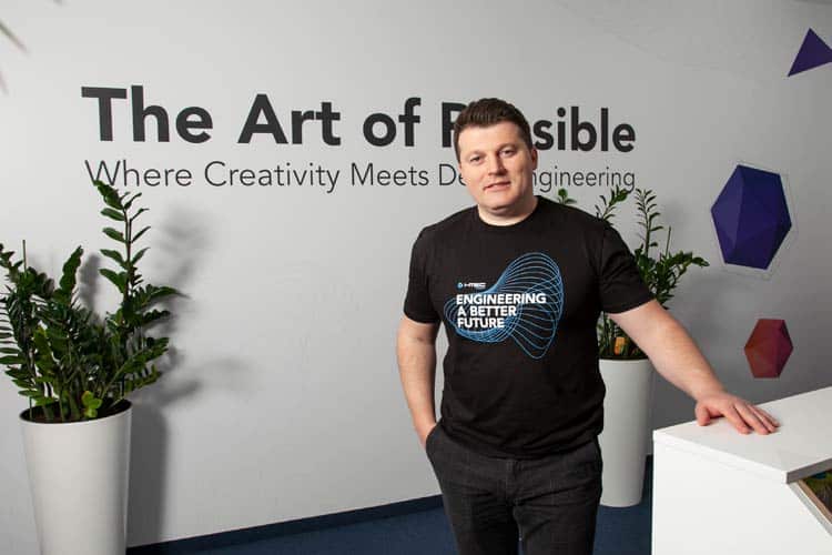 A man in a black T-shirt that reads "Engineering a Better Future" stands next to a white wall with the text "The Art of Possible: Where Creativity Meets Engineering" on it. There are two potted plants on either side of him and geometric wall decorations in the background.