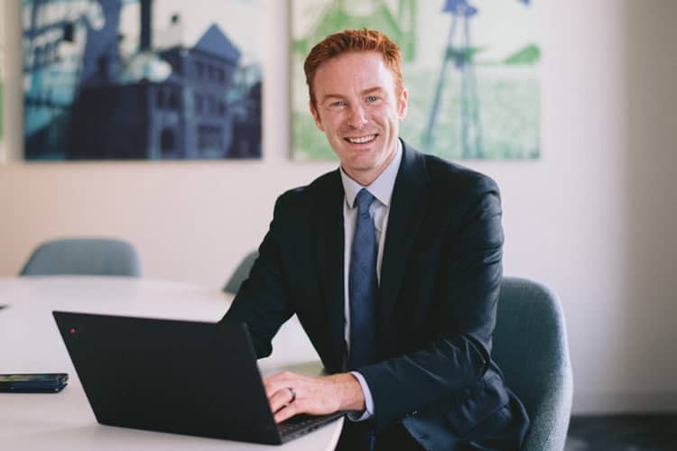 A man wearing a dark blue suit and tie is seated at a desk with a laptop in front of him. He is smiling and looking at the camera. In the background, there are abstract art pieces on the wall. The setting appears to be a modern office or conference room.