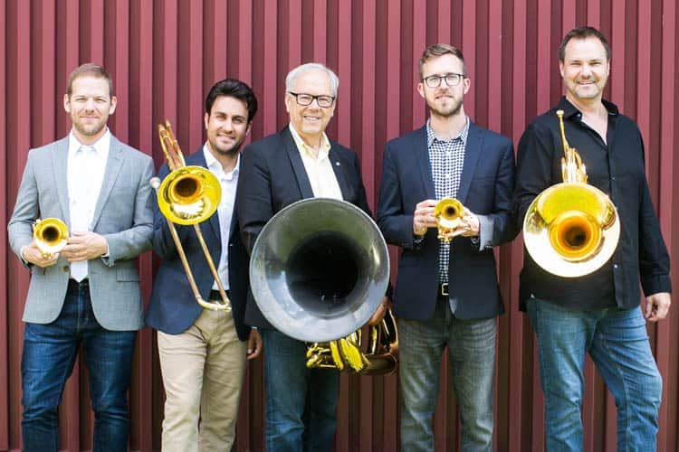 Five men stand in front of a vertical red-striped wall, each holding brass instruments, including trombones and a tuba. Four men wear blazers with shirts, one wears a black shirt without a blazer. They all smile at the camera.
