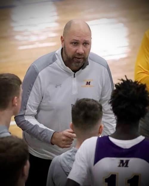 A coach with a bald head and beard, wearing a white and gray "M" logo jacket, gives instructions to a group of basketball players in a gym. The players are huddled around, listening intently, with one player wearing a purple and white jersey numbered 11 in the foreground.