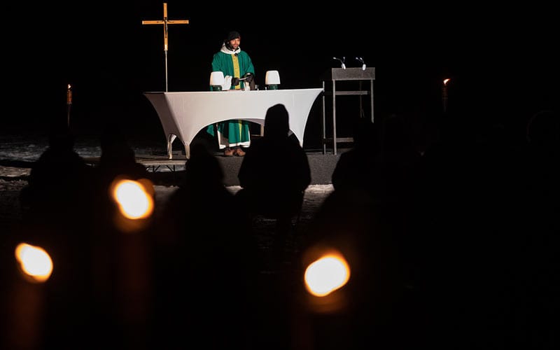 A priest dressed in green stands behind a white altar, illuminated by a spotlight. The scene is dark, with dimly lit worshippers in the foreground and a golden cross in the background. Candlelights create a serene atmosphere.