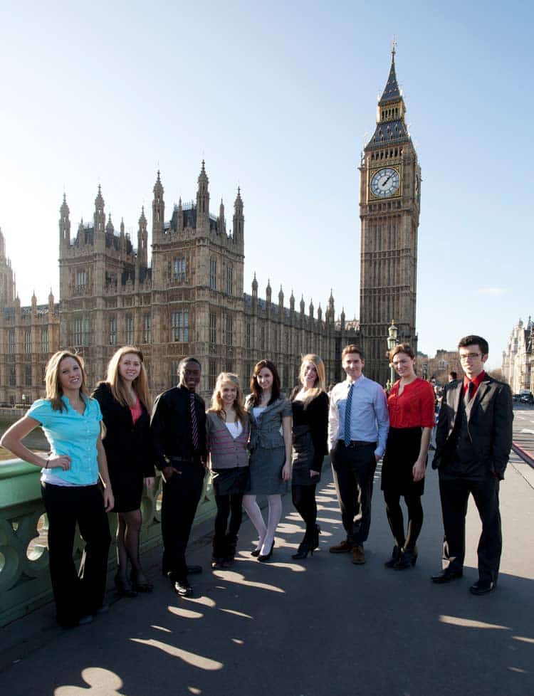 A group of nine people, both men and women, stand on a bridge with the Palace of Westminster and Big Ben in the background. They are dressed in business attire and smile at the camera on a sunny day.