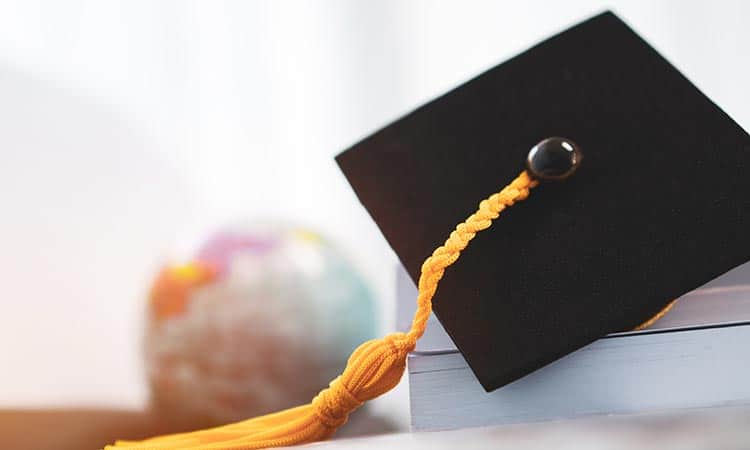 A close-up of a black graduation cap with a yellow tassel resting on a stack of books. A blurred miniature globe is visible in the background, suggesting themes of education and global learning. The scene is well-lit with a soft focus on the background.