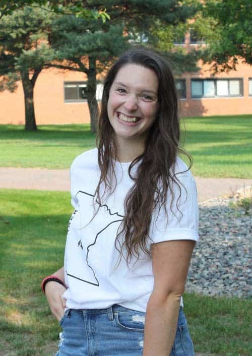 A woman with long, wavy hair smiling at the camera. She is wearing a white t-shirt with an outline drawing on it and denim shorts. The background features grass, a gravel path, trees, and a brick building with windows.