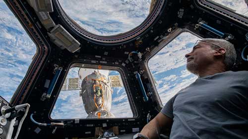 An astronaut is looking out of a cupola module at the International Space Station. The view through the windows shows the Earth with clouds below and another part of the space station clearly visible. The image shares a sense of the serenity and vastness of space.