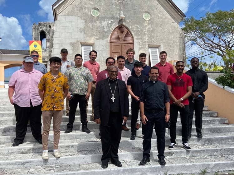 A group of people, including clergymen and young men, stand on the stone steps in front of an old church building. The sky is partly cloudy. The church has a wooden door and a small arched window above it. Everyone is dressed in casual, semi-formal attire.