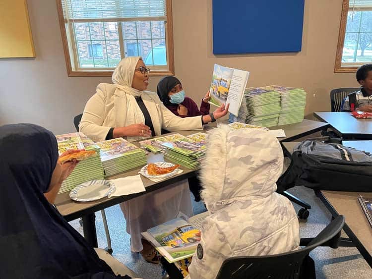 A woman in a headscarf reads a book to three children seated around a table. The table is covered with stacks of books and some pizza. The children appear engaged, and one of them is wearing a face mask. The room has windows, blue wall panels, and other students in the background.
