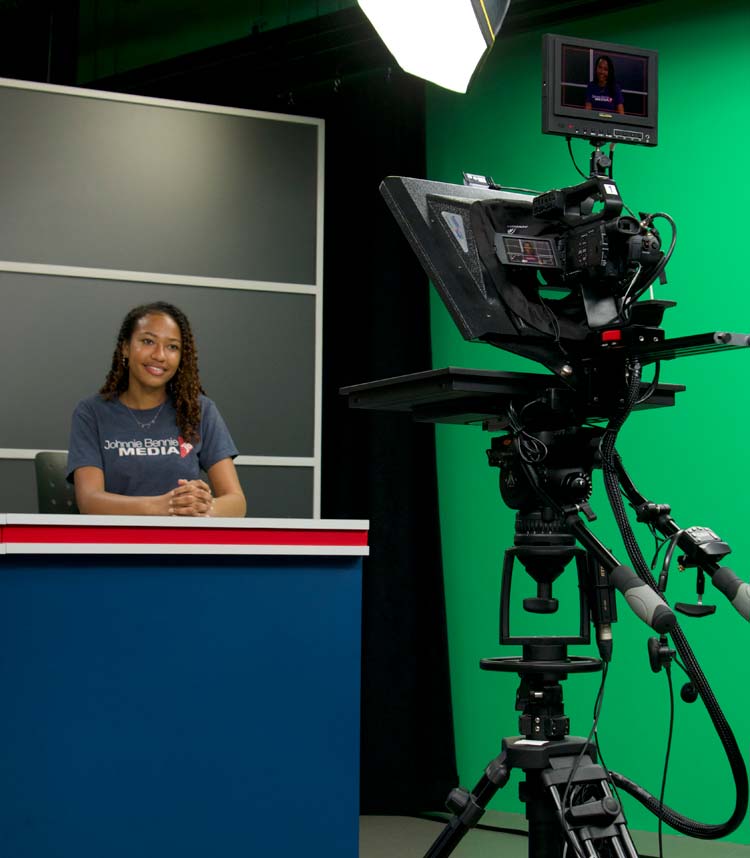 A person sits at a desk in a studio setting with green screen and professional video camera equipment. The individual has long hair, is smiling, and wears a t-shirt. The studio lighting is bright, and there's a screen displaying their image on top of the camera.