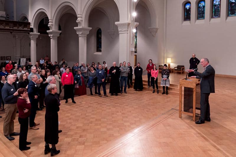 A speaker addresses a moderately sized audience in an ornate hall with high arches and stained glass windows. The attendees, varying in age and attire, attentively listen while standing on the wooden floor. A microphone and lectern are visible.