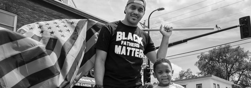 A man and a young boy stand together outdoors, the man holding an American flag on a pole. The man is wearing a shirt that reads "BLACK FATHERS MATTER." They appear to be participating in a community or social event, with buildings and a traffic light visible in the background.