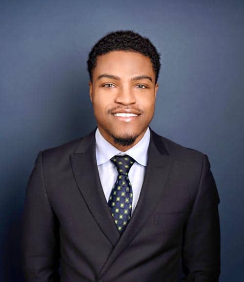 A man with short curly hair and a trimmed beard is smiling. He is wearing a dark suit jacket, a light blue dress shirt, and a checkered tie. The background is a plain, dark blue-gray color.