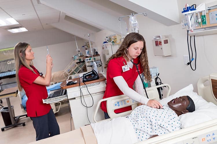 Two healthcare professionals in red scrubs practice medical procedures in a simulation lab. One adjusts an IV drip, while the other checks the breathing of a medical mannequin on a hospital bed, surrounded by medical equipment.