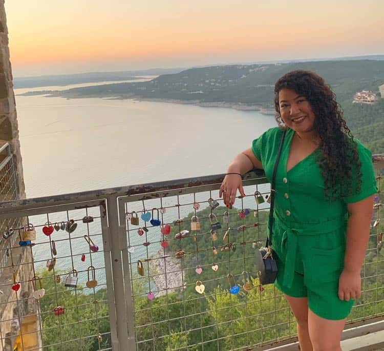 Woman in a green dress standing by a fence adorned with love locks, with a picturesque lake and mountainous landscape in the background during sunset. She is smiling at the camera and holding onto the railing.