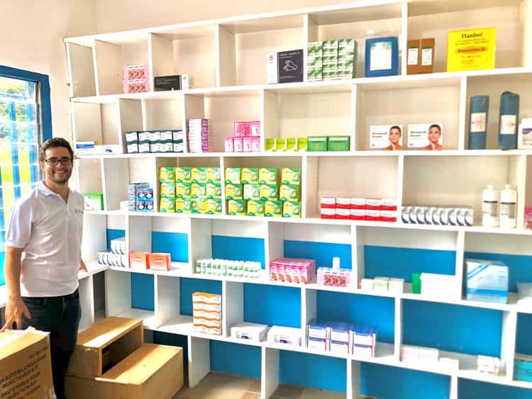 A smiling man in a white shirt stands next to white shelves filled with various medical and skincare products, arranged neatly in boxes and bottles. Cardboard boxes are on the floor nearby. The shelves are mostly empty with some items in select compartments.