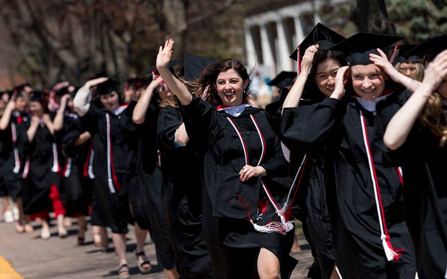 A group of graduates in black gowns and caps, adorned with red and white tassels, are joyfully walking outdoors. Several are waving and smiling, celebrating their achievement. Trees and a building with white columns are visible in the background.
