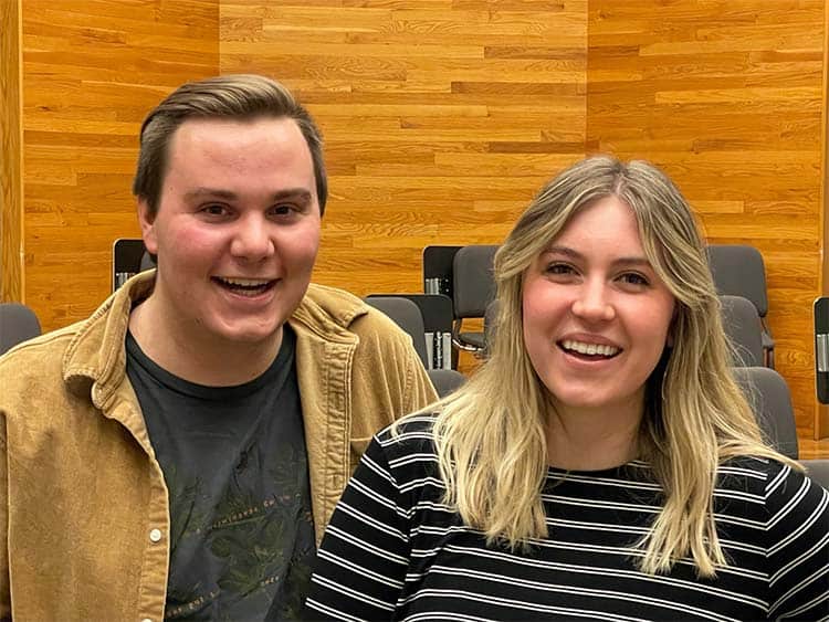 Two people smiling in a room with wooden walls and rows of empty chairs behind them. The person on the left has short hair and wears a light brown jacket over a dark shirt. The person on the right has long, light hair and wears a black and white striped shirt.