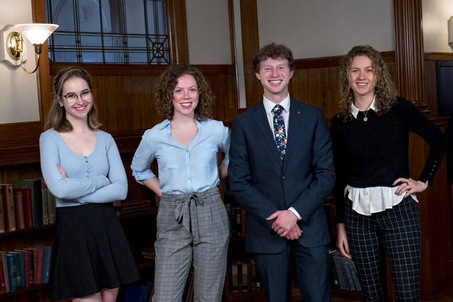 Four people are standing in a room with wood-paneled walls and a bookshelf. From left to right: a person in glasses, a light blue top, and a black skirt; a person in a light blue blouse and checkered pants; a person in a suit and tie; and a person in black top and striped pants.